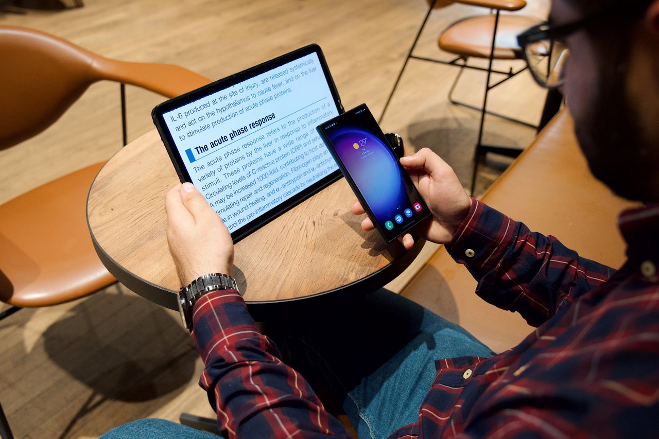 about-img A man using a tablet and smartphone at a coffee table in a modern indoor setting.