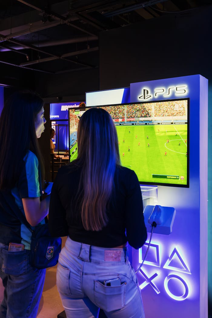 about-img Two women playing FIFA on a PlayStation 5 console in a modern gaming store.